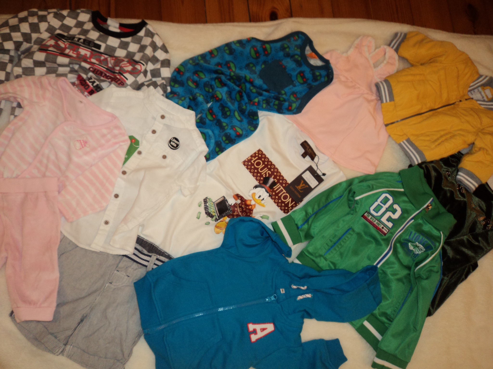 Rozłożona kolekcja różnorodnych ubrań dziecięcych, w tym bluza z kapturem, koszulka z nadrukiem, spodnie dresowe, kurtka bomberka i koszula z krótkim rękawem, ułożone na białym tle.