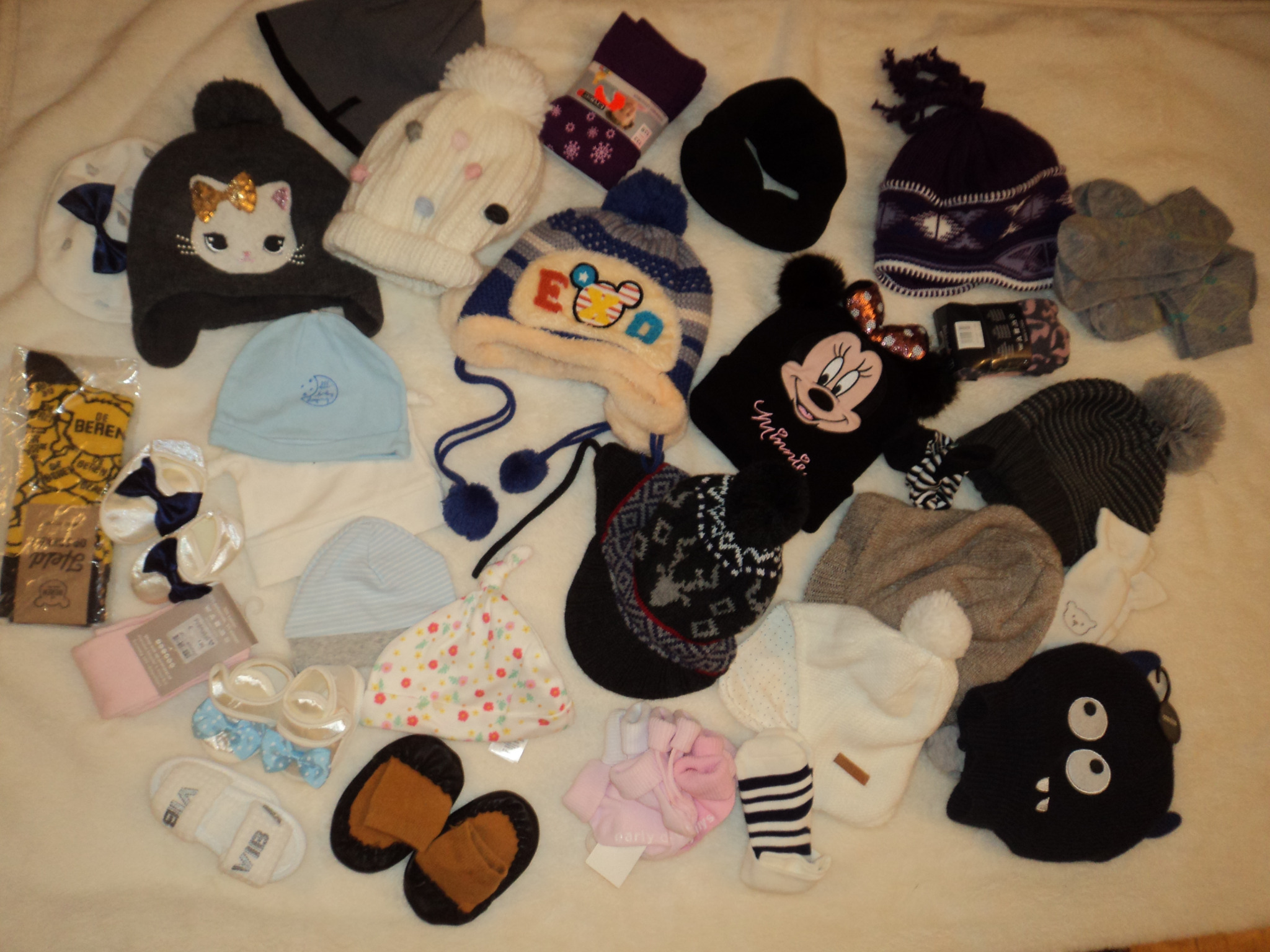 Kolekcja czapek dziecięcych, skarpetek i bucików na białym, puszystym tle. Widoczne czapki z motywami zwierzęcymi, pomponami i wzorami, obok nich skarpetki w różnych kolorach i wzorach oraz małe...