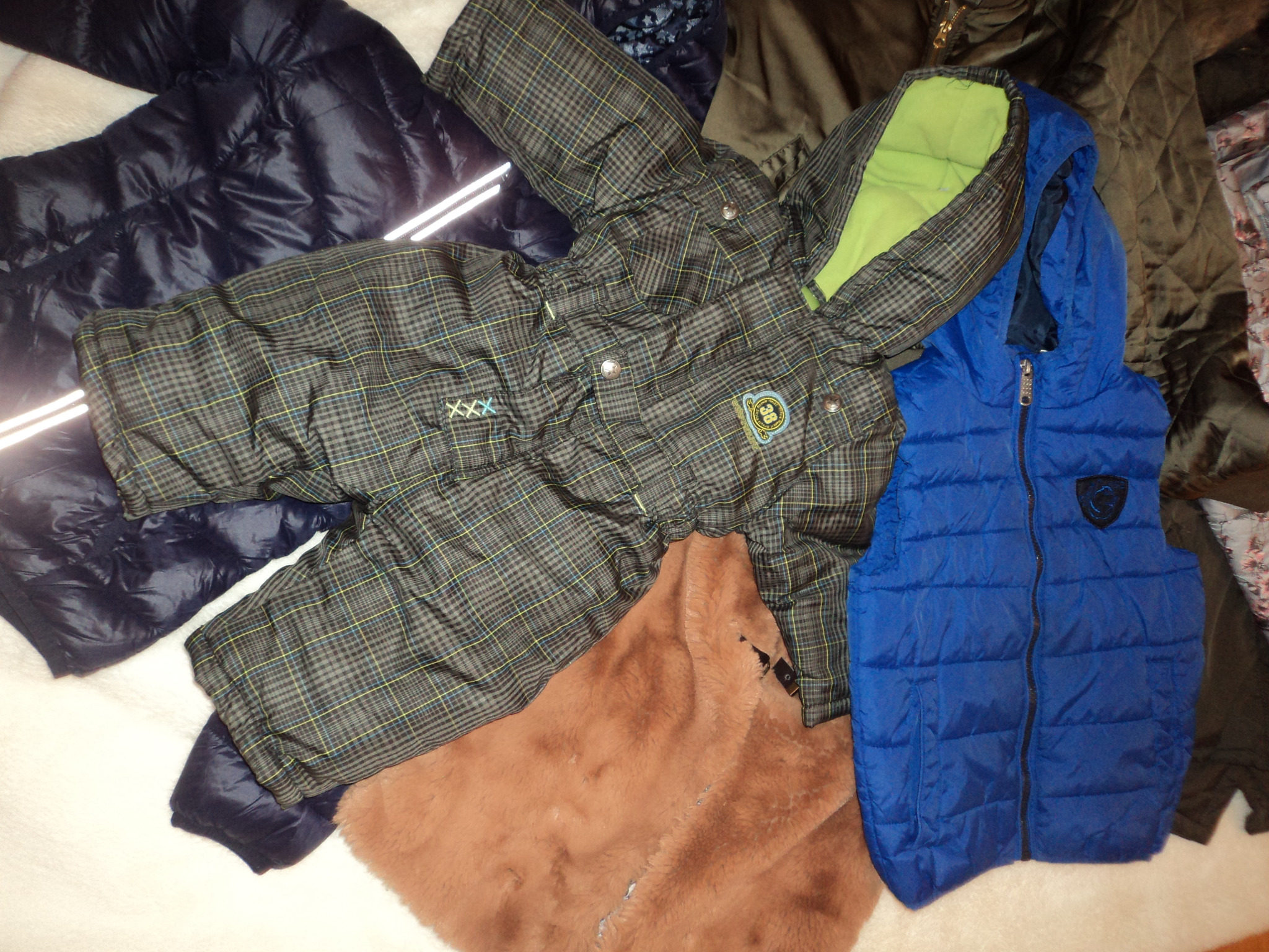 Stos ubrań: granatowa kurtka puchowa z odblaskami, kombinezon w kratę z zieloną podszewką, niebieska kamizelka puchowa i brązowe futro.