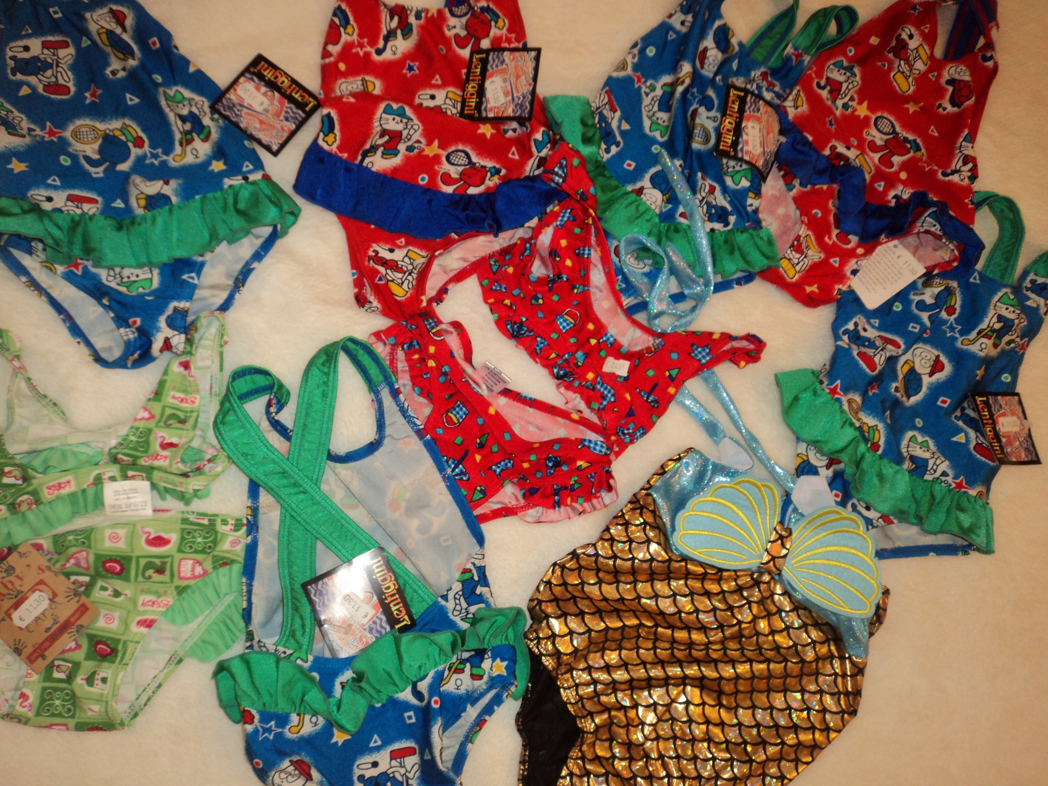 Różnokolorowe stroje kąpielowe dziecięce z wzorami, niektóre z falbankami i metkami, ułożone na białym tle.