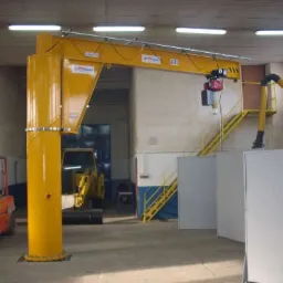 Żółty żuraw warsztatowy stacjonarny z wciągarką łańcuchową w hali produkcyjnej, z widocznym w tle wózkiem widłowym i koparką.