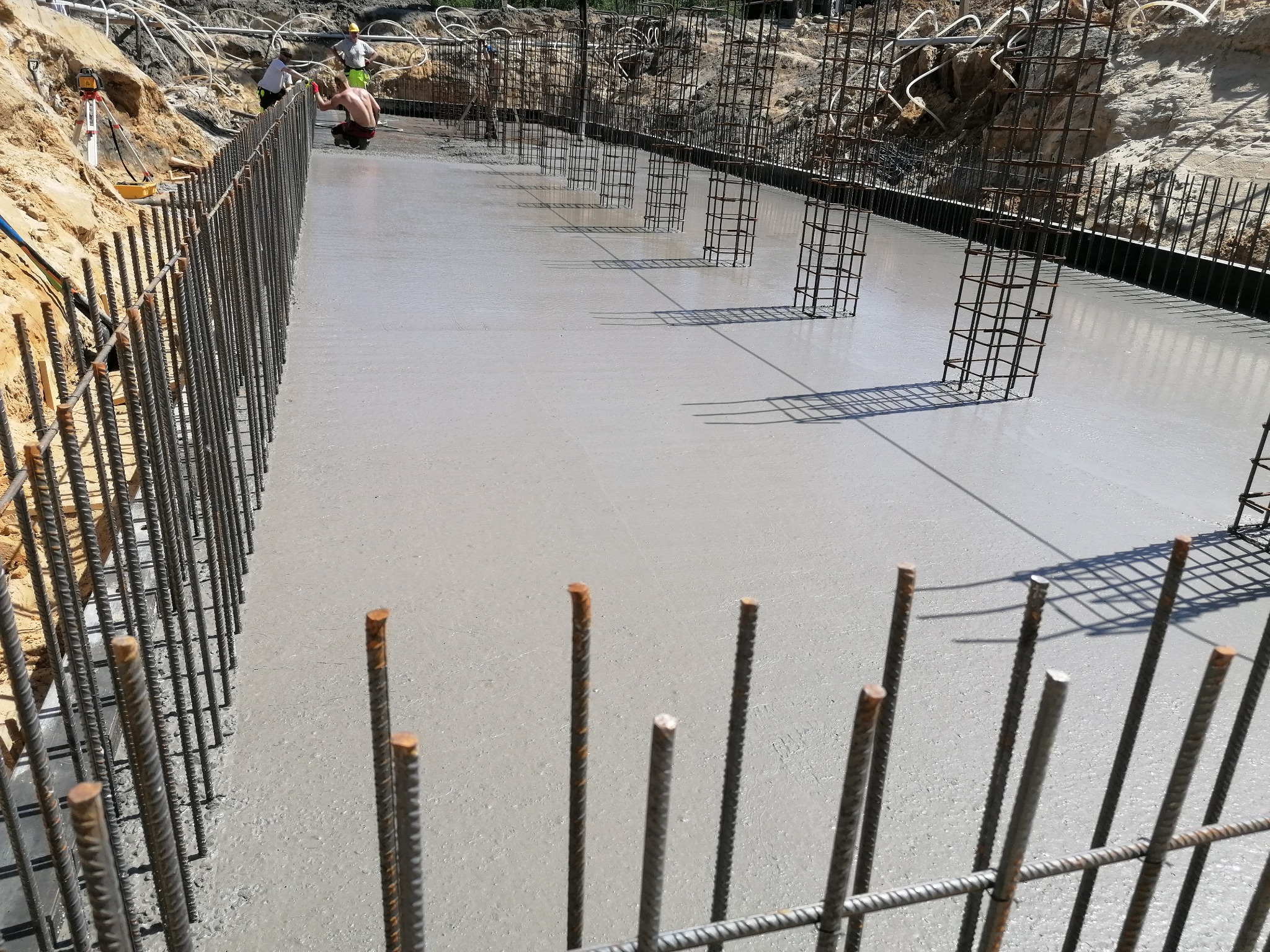 Świeżo wylany beton w szalunku zbrojonym stalowymi prętami, widoczni pracownicy budowlani w tle, teren budowy w wykopie.
