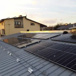 Panele słoneczne zamontowane na metalowym dachu domu jednorodzinnego, widoczne elementy montażowe i fragmenty innych instalacji na dachu sąsiedniego budynku.