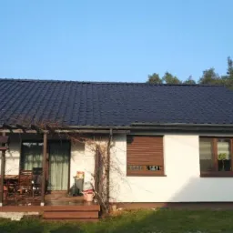 Dom jednorodzinny z ciemną dachówką modułową i pergolą porośniętą winoroślą, widok z ogrodu w słoneczny dzień.