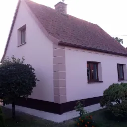 Jasny, otynkowany dom jednorodzinny z czerwoną dachówką, widok z boku na elewację z trzema oknami i ozdobnymi narożnikami, otoczony zielenią.