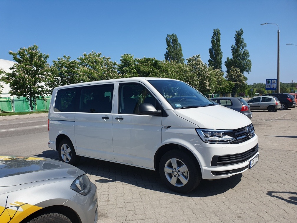 Biały van Volkswagen Transporter T6 na parkingu w słoneczny dzień, widoczne drzewa i inne samochody w tle.