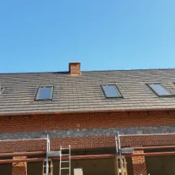 Montaż okna dachowego w nowym, brązowym dachu z dachówki ceramicznej, widoczne rusztowania i fragment murowanej ściany z cegły klinkierowej.