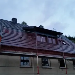 Dom w trakcie prac dekarskich, z nowym, ciemnym pokryciem dachowym i rusztowaniem ochronnym, widoczne okno dachowe i komin.