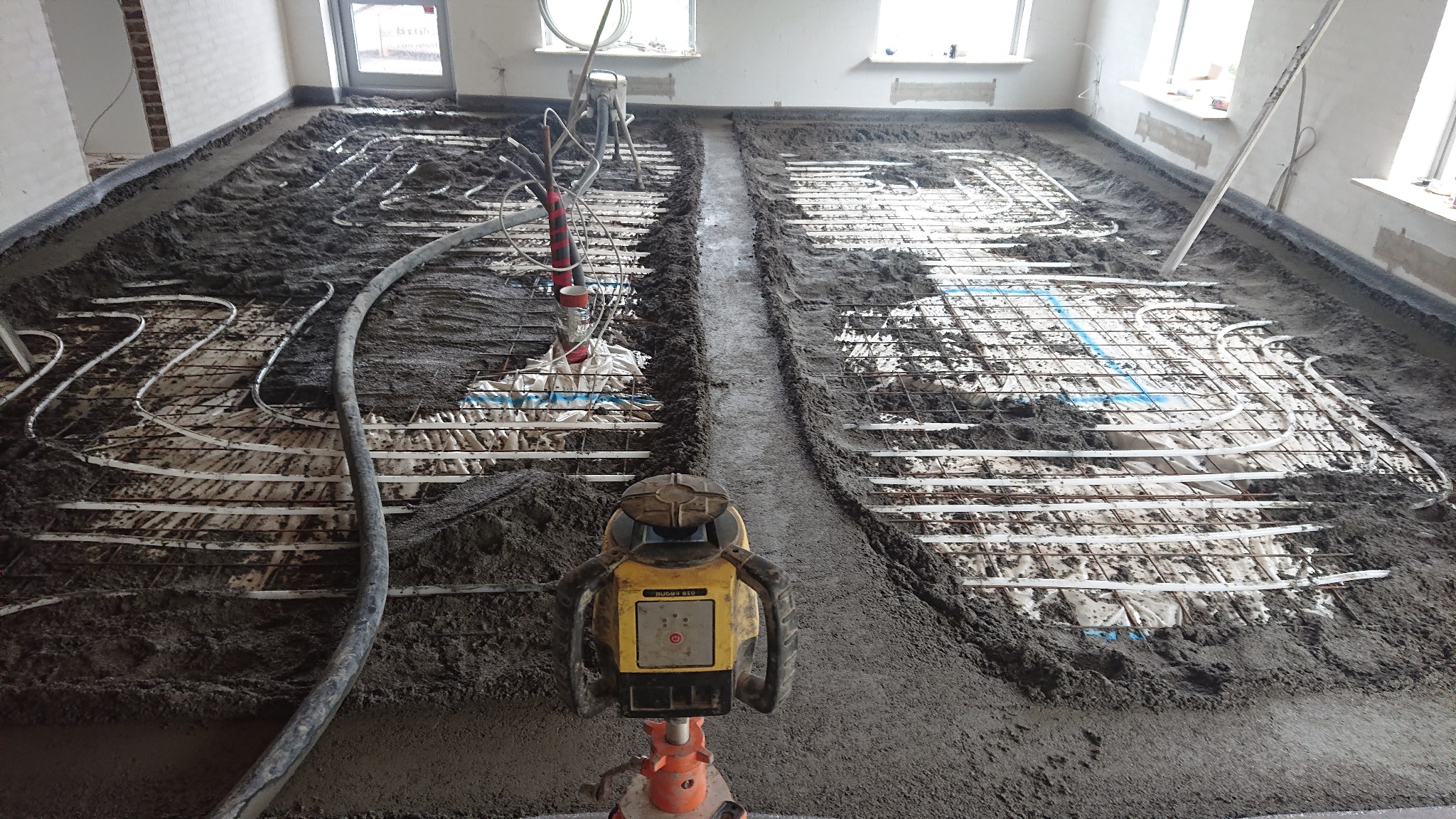 Wnętrze w trakcie wylewania posadzki z ogrzewaniem podłogowym, widoczna siatka z rurami grzewczymi zalewana mokrą zaprawą, poziomica laserowa ustawiona na trójnogu.