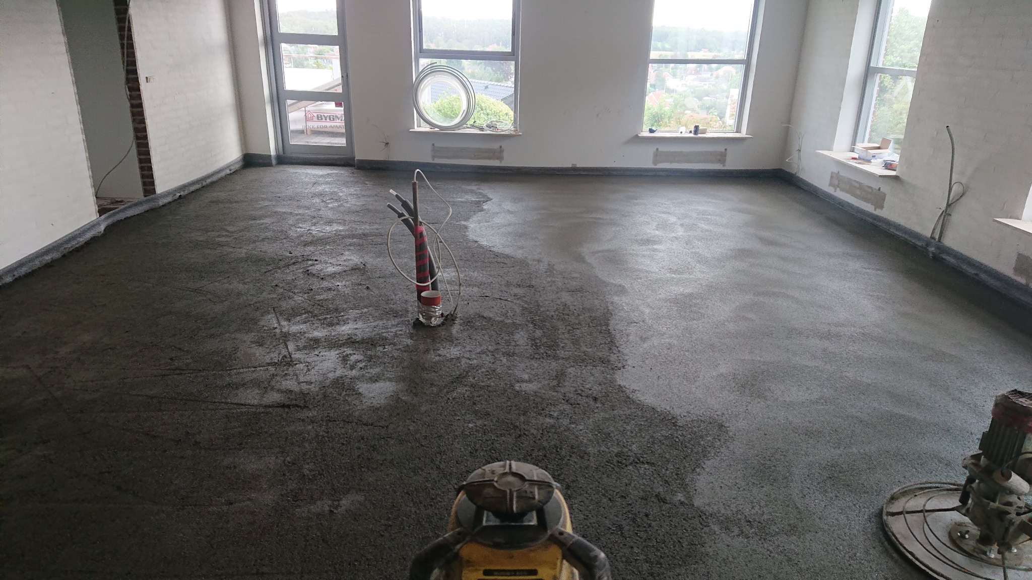 Wnętrze w trakcie wylewania posadzki jastrychowej, widoczne okna, grzejniki i maszyny do wygładzania betonu.