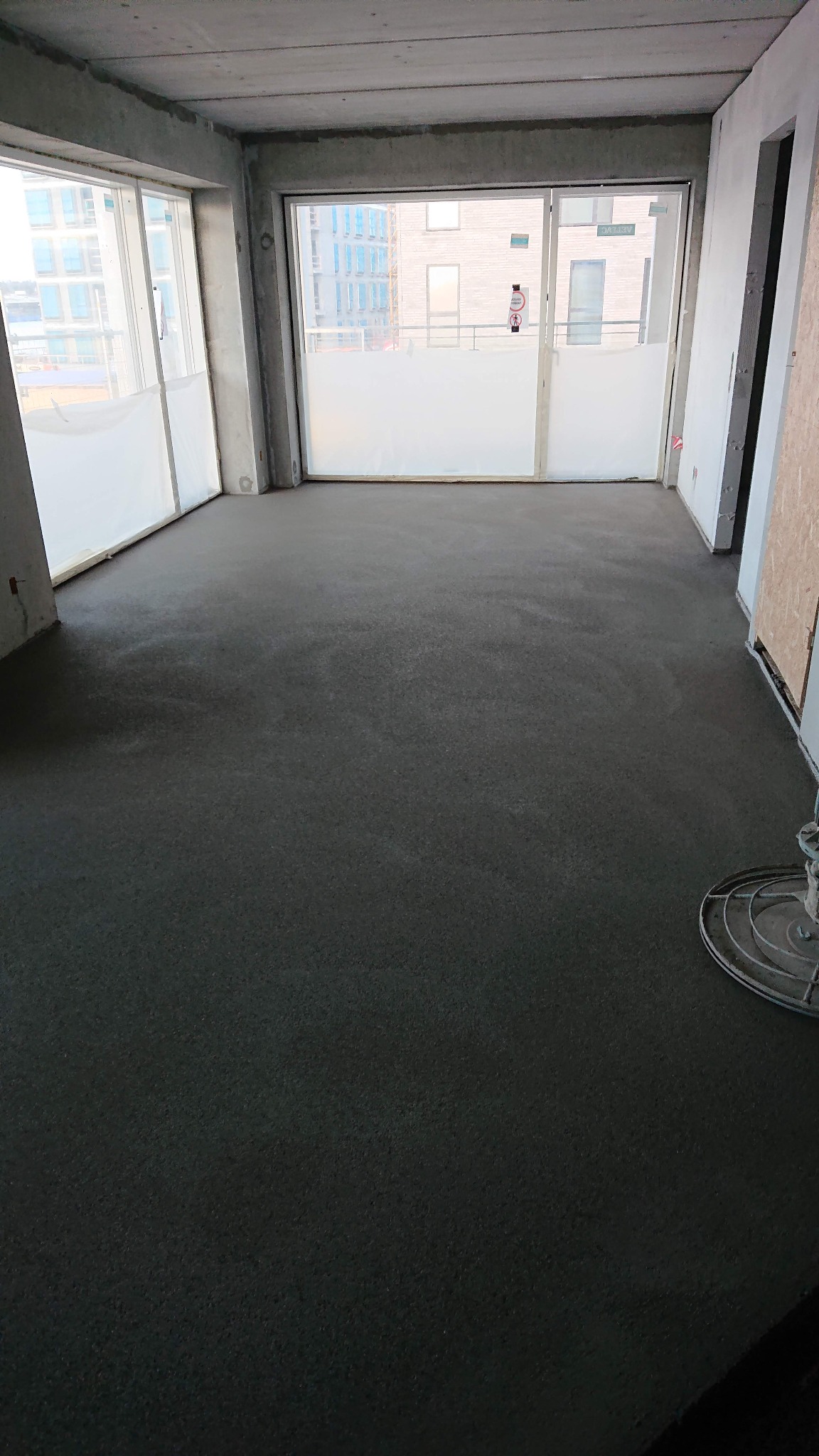 Świeżo wylana, ciemna posadzka w surowym wnętrzu z dużymi oknami, widoczna maszyna do zacierania posadzek w prawym dolnym rogu.