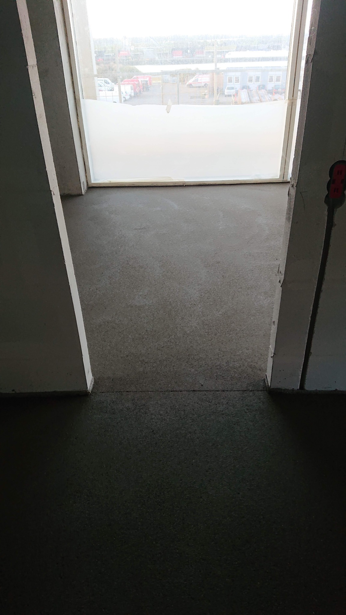 Świeżo wylana, gładka posadzka w surowym wnętrzu, widok przez otwór okienny z zamazanym widokiem zewnętrznym.