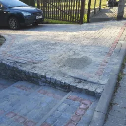 Ułożona kostka brukowa z dekoracyjnym obramowaniem, widoczne schody z kostki, mały kopiec piasku na powierzchni, samochód osobowy w tle, drewniany płot po lewej stronie.