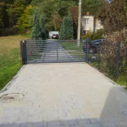 Nowoczesna posesja z szarą kostką brukową prowadzącą do metalowej bramy i ogrodzenia panelowego, w tle zieleń drzew i dom.