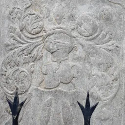 Szara kamienna płaskorzeźba przedstawiająca rycerza w hełmie otoczonego ornamentami roślinnymi, widziana przez fragment metalowego ogrodzenia.