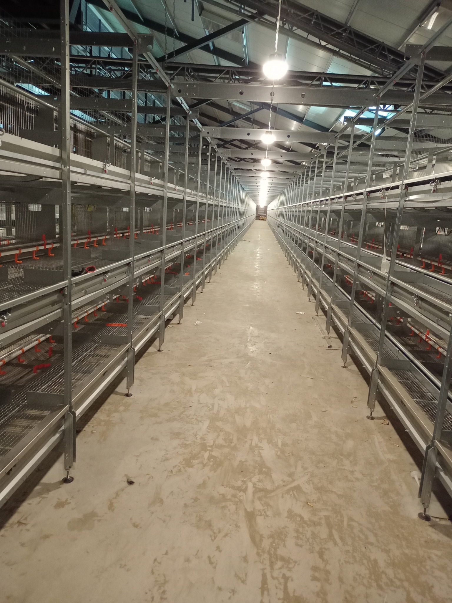 Długi korytarz z metalowymi konstrukcjami regałowymi, oświetlony lampami sufitowymi, posadzka betonowa z widocznymi śladami.