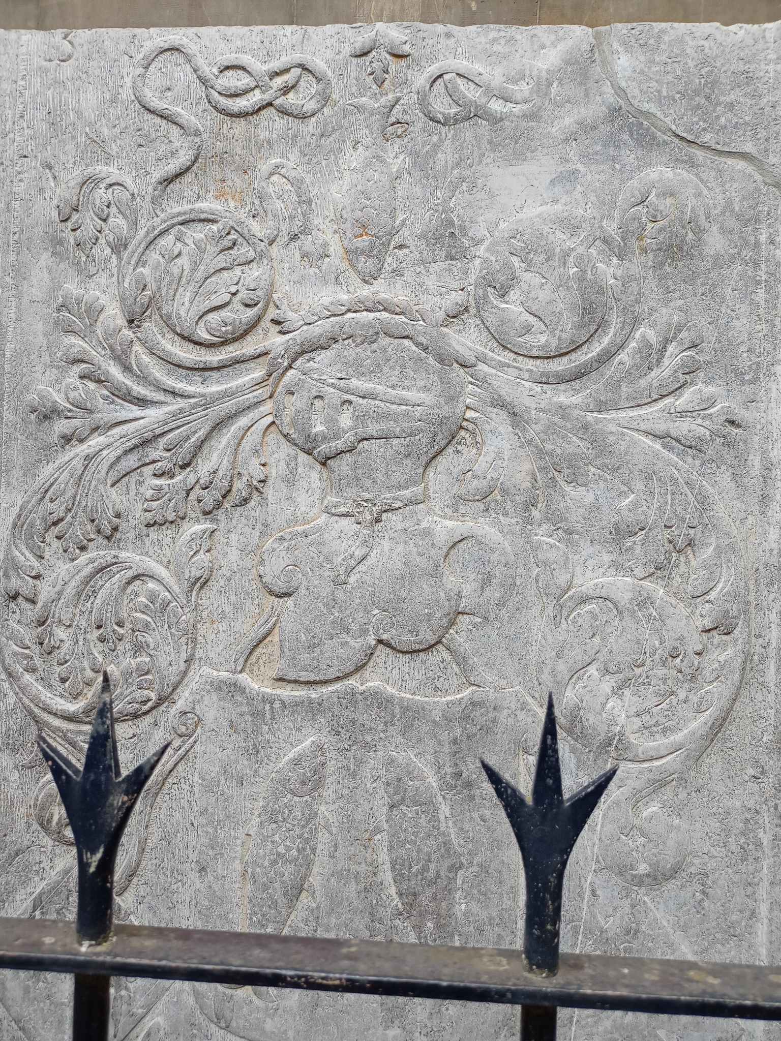 Szara kamienna płaskorzeźba przedstawiająca rycerza w hełmie otoczonego ornamentami roślinnymi, widziana przez fragment metalowego ogrodzenia.