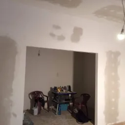 Pokój w trakcie szpachlowania ścian, widoczne miejsca po łataniu ubytków, prowizoryczne oświetlenie żarówką na kablu, stolik z narzędziami i napojami, dwa plastikowe krzesła.