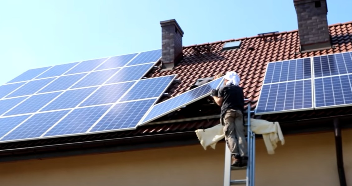 Montaż paneli słonecznych na dachu domu z ceglaną dachówką, instalator na drabinie w trakcie pracy.
