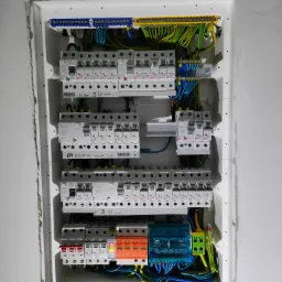 Rozdzielnica elektryczna z bezpiecznikami, zabezpieczeniami przepięciowymi i listwami zaciskowymi, widoczne przewody w różnych kolorach, zamontowana w ścianie.