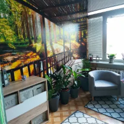 Gabinet z fototapetą leśnego krajobrazu, szarym fotelem i roślinami doniczkowymi, tworzący spokojną atmosferę.