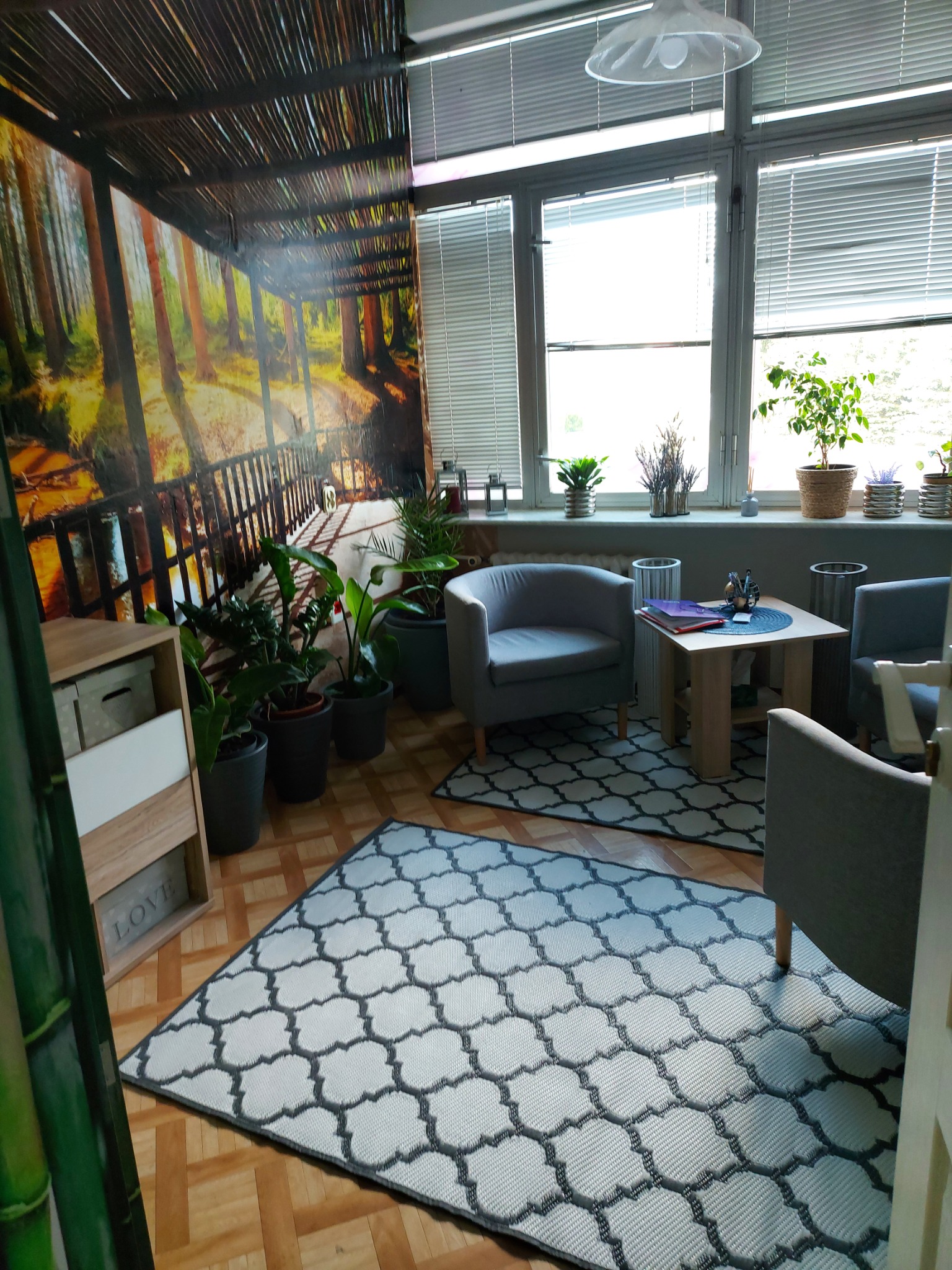 Jasne wnętrze gabinetu z fototapetą leśną, dwoma szarymi fotelami, stolikiem z przyborami biurowymi i dywanem w geometryczne wzory.