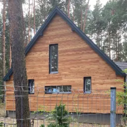 Nowoczesny dom jednorodzinny z elewacją z jasnego drewna, czarnym dachem i ramami okiennymi, otoczony lasem, widoczny zza metalowego ogrodzenia.