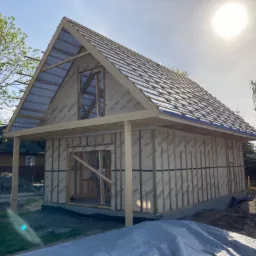 Budowa szkieletu domu jednorodzinnego w trakcie montażu dachu, widoczne membrany dachowe z nadrukiem producenta, drewniana konstrukcja ścian i zadaszenia, teren budowy z ziemią i plandeką.