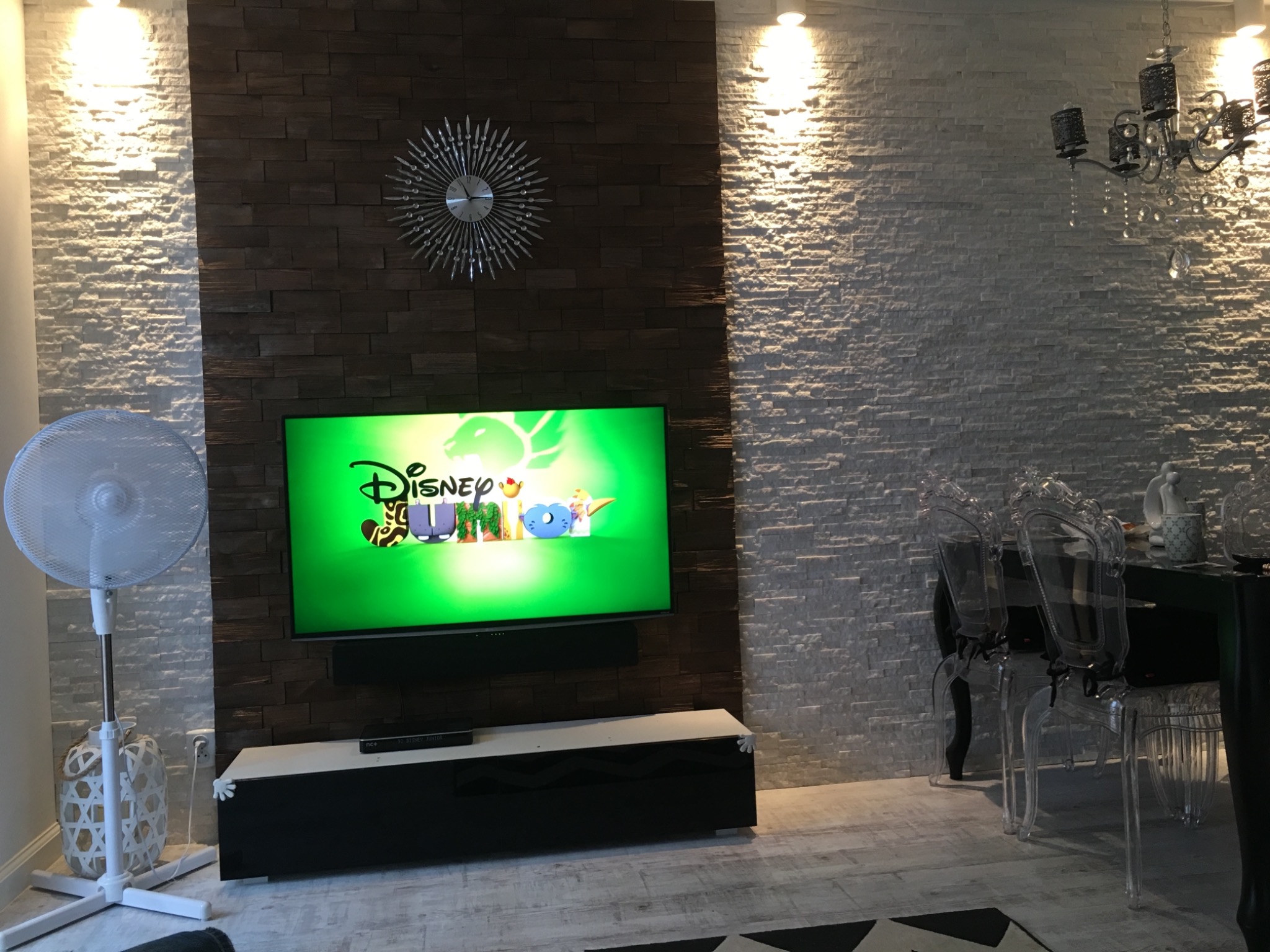 Salon z dekoracyjną ścianą z imitacji kamienia w dwóch odcieniach, telewizorem z włączoną bajką, ozdobnym zegarem i designerskim żyrandolem
