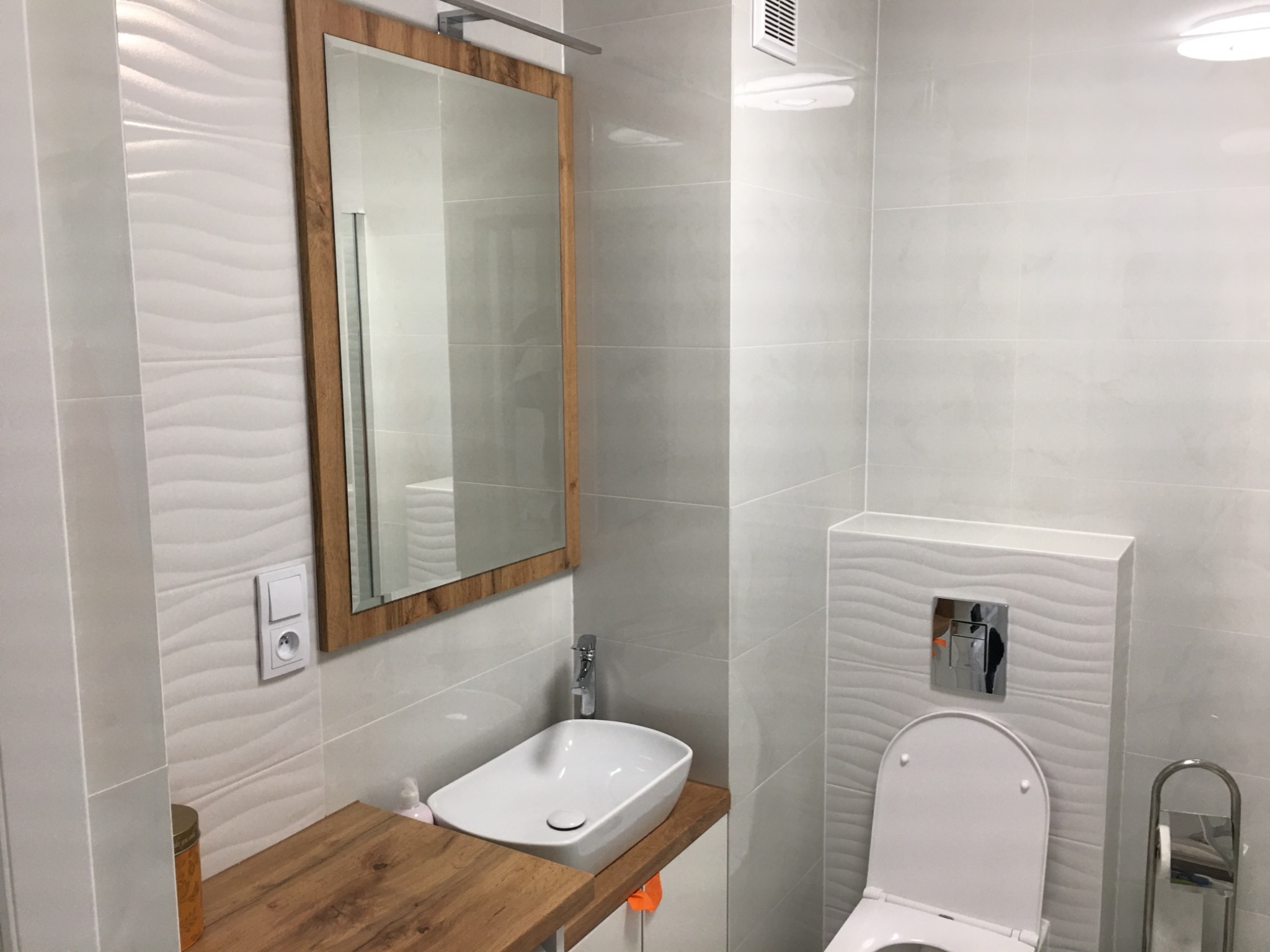 Nowoczesna łazienka z umywalką nablatową na drewnianym blacie, lustrem w drewnianej ramie i toaletą z podtynkową spłuczką, wykończona białymi płytkami z falistym wzorem.