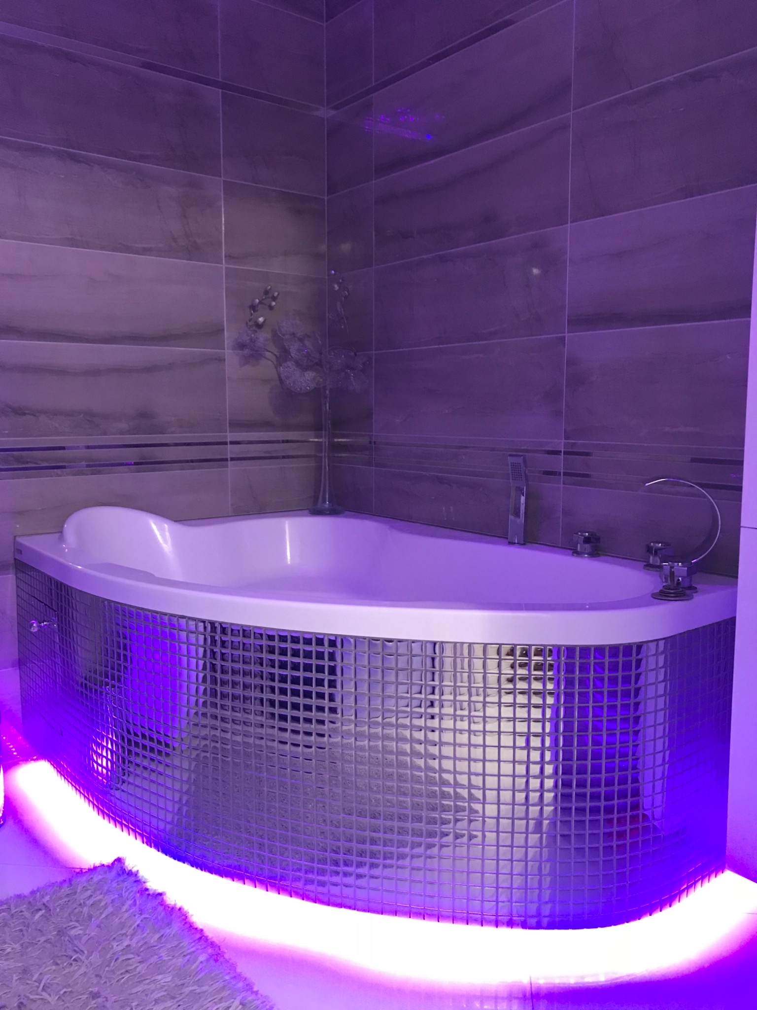 Nowoczesna wanna narożna z mozaikową obudową podświetloną fioletowym światłem LED, szare płytki na ścianach, dekoracyjny wazon z kwiatami.