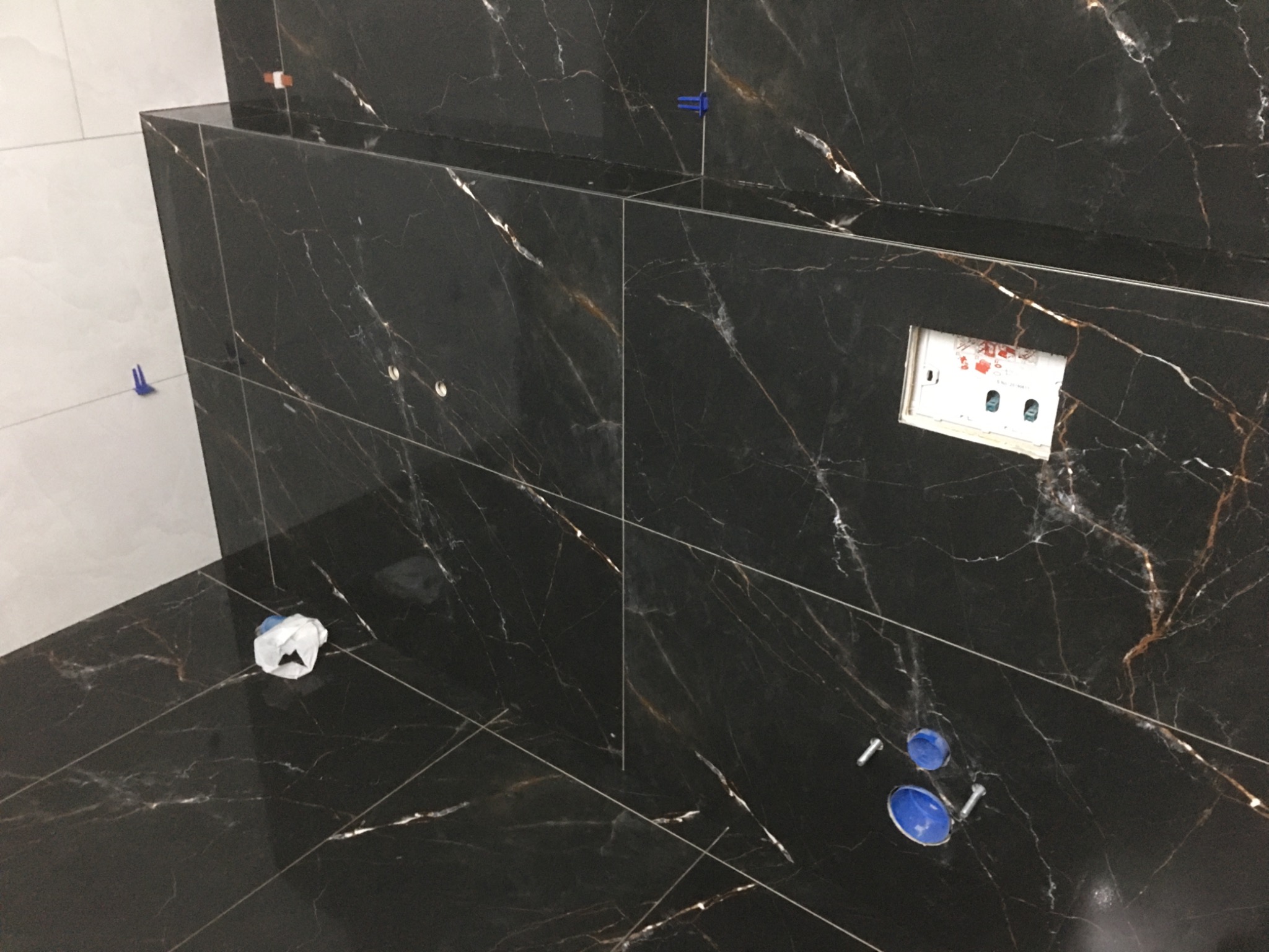 Ułożone płytki imitujące czarny marmur z białymi i złotymi żyłami na ścianie i podłodze, widoczne plastikowe krzyżyki dystansowe, puszka elektryczna i elementy instalacji sanitarnej.
