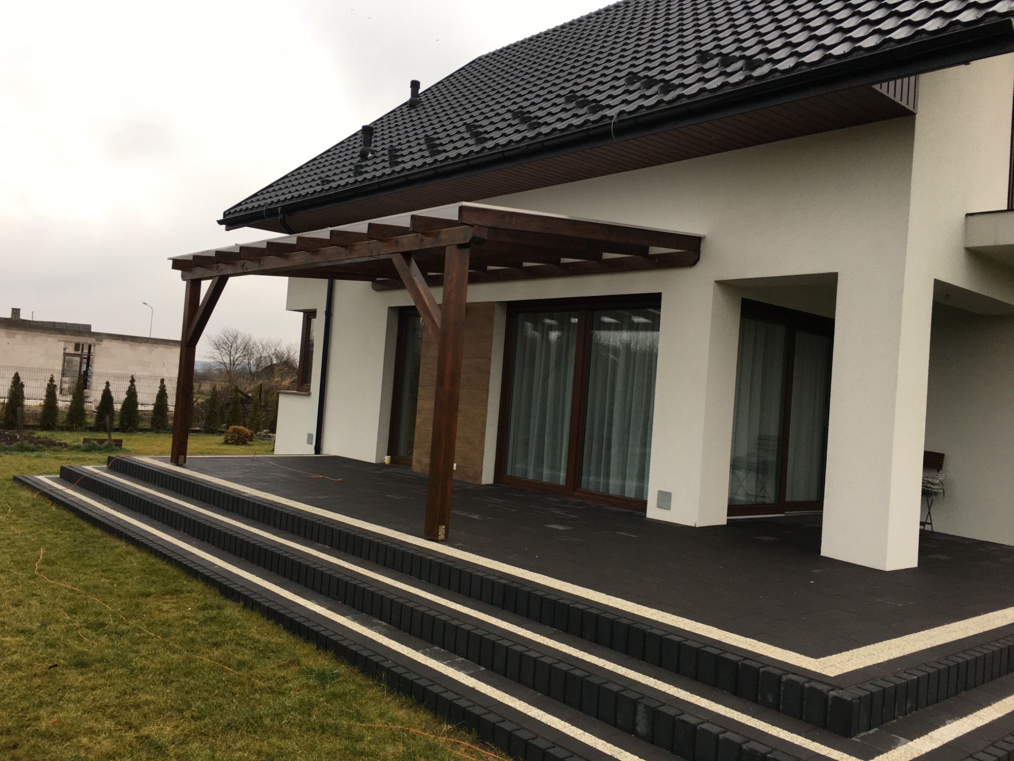 Nowoczesny dom z ciemną dachówką i drewnianą pergolą nad tarasem wyłożonym ciemną kostką z jasnymi akcentami, widoczne schody z kostki.
