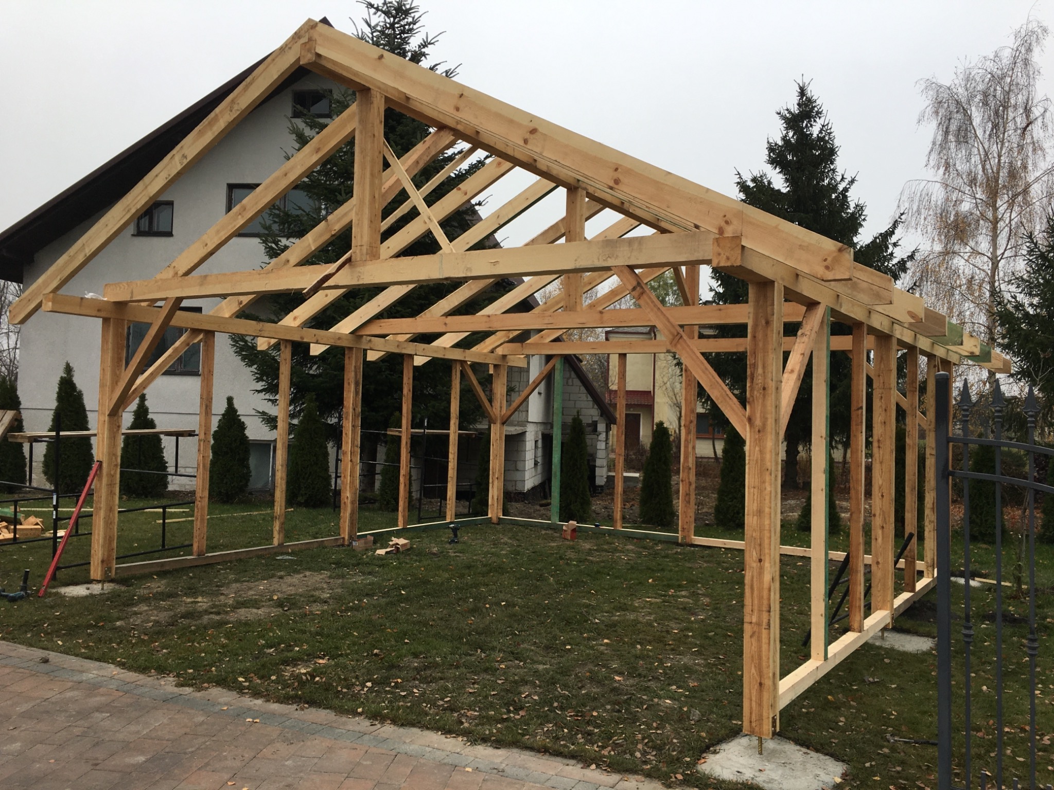 Konstrukcja drewniana z belek i słupów, prawdopodobnie wiata garażowa w trakcie budowy, widoczna na tle domu jednorodzinnego i zieleni.
