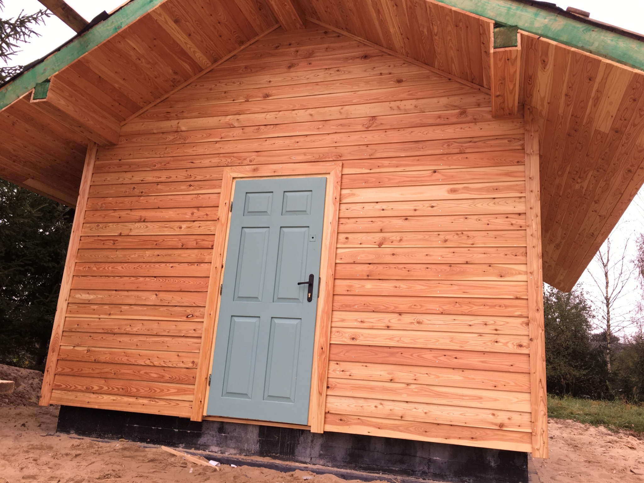 Drewniana elewacja budynku z deskami ułożonymi poziomo, szara, panelowa brama wejściowa z czarną klamką, widoczne elementy konstrukcji dachu.