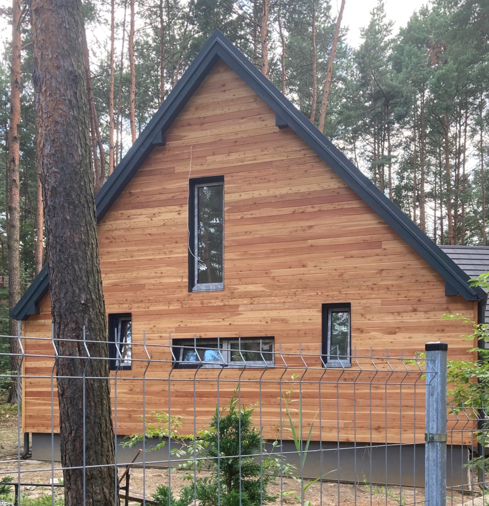Nowoczesny dom jednorodzinny z elewacją z jasnego drewna, czarnym dachem i ramami okiennymi, otoczony lasem, widoczny zza metalowego ogrodzenia.