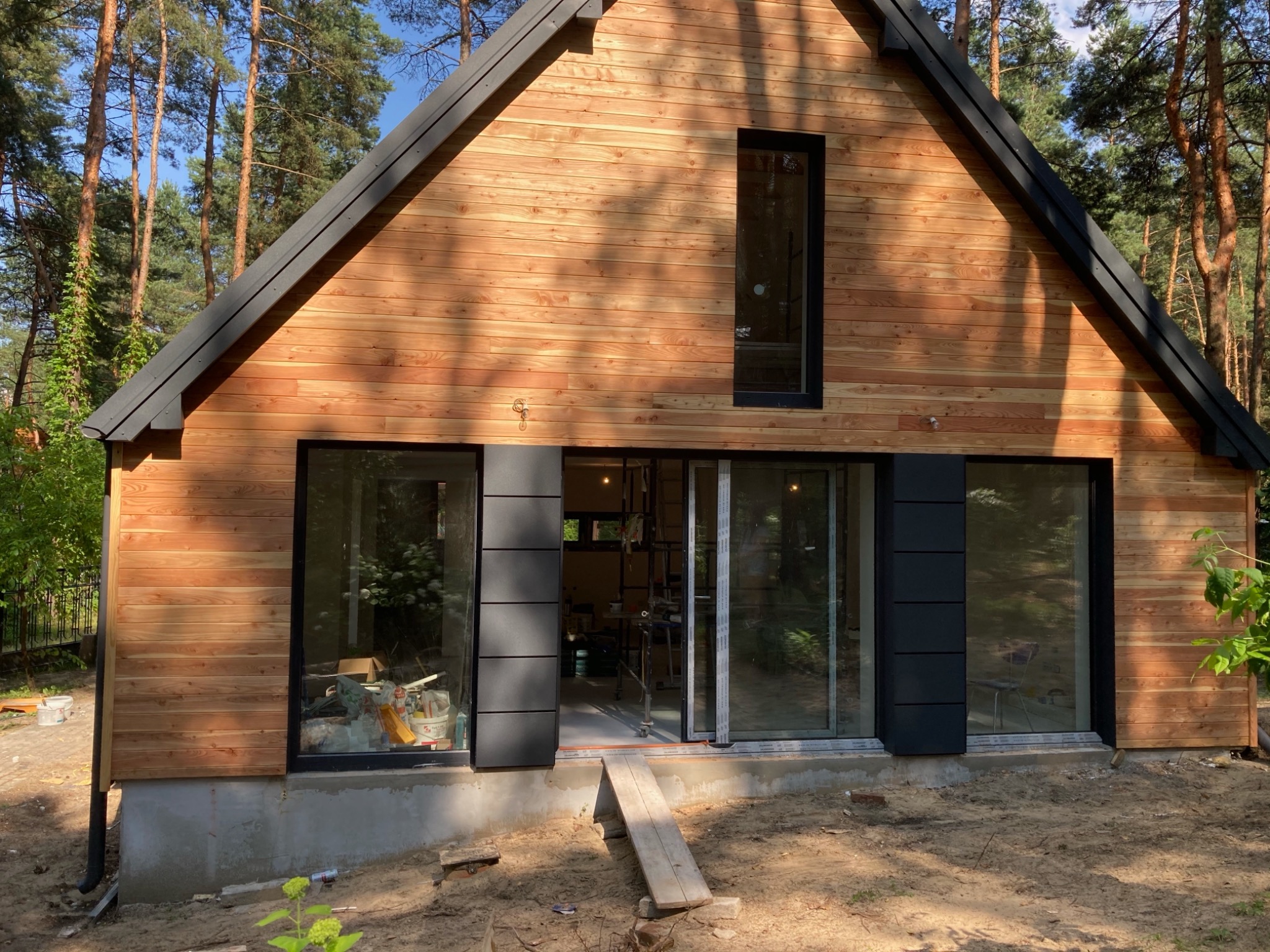 Nowoczesny dom jednorodzinny z drewnianą elewacją i czarnymi ramami okiennymi, widoczne rusztowania wewnątrz, teren wokół domu w trakcie zagospodarowywania.