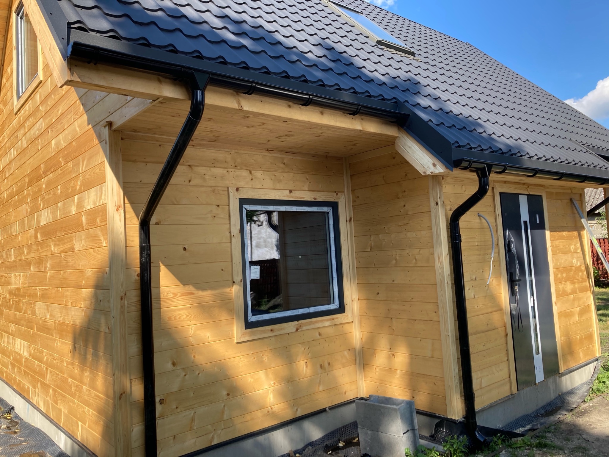 Nowoczesny, drewniany dom z czarnym dachem z blachodachówki i systemem rynnowym w kolorze czarnym, okna w czarnych ramach, jasne drewniane deski elewacyjne.