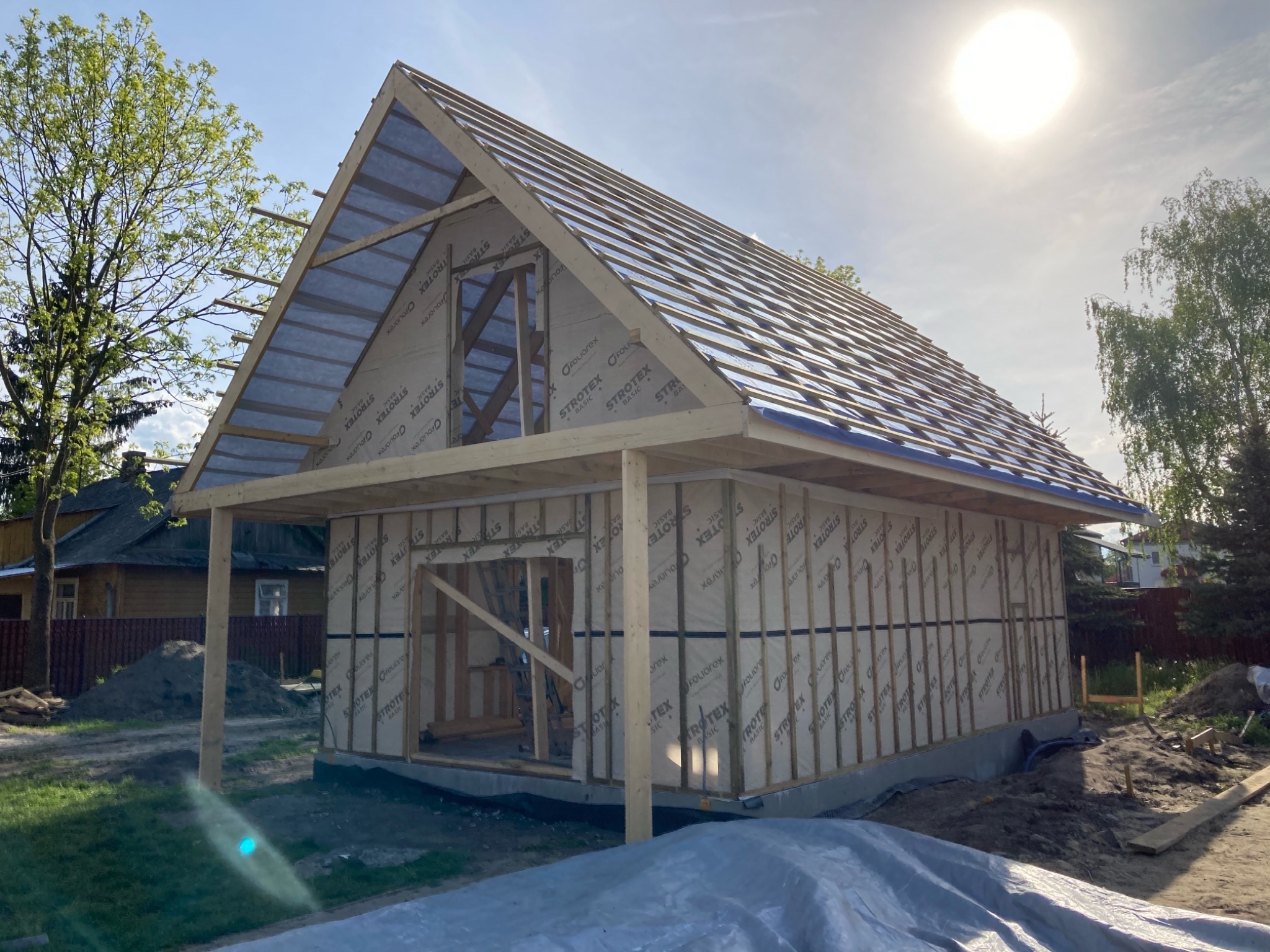 Budowa szkieletu domu jednorodzinnego w trakcie montażu dachu, widoczne membrany dachowe z nadrukiem producenta, drewniana konstrukcja ścian i zadaszenia, teren budowy z ziemią i plandeką.