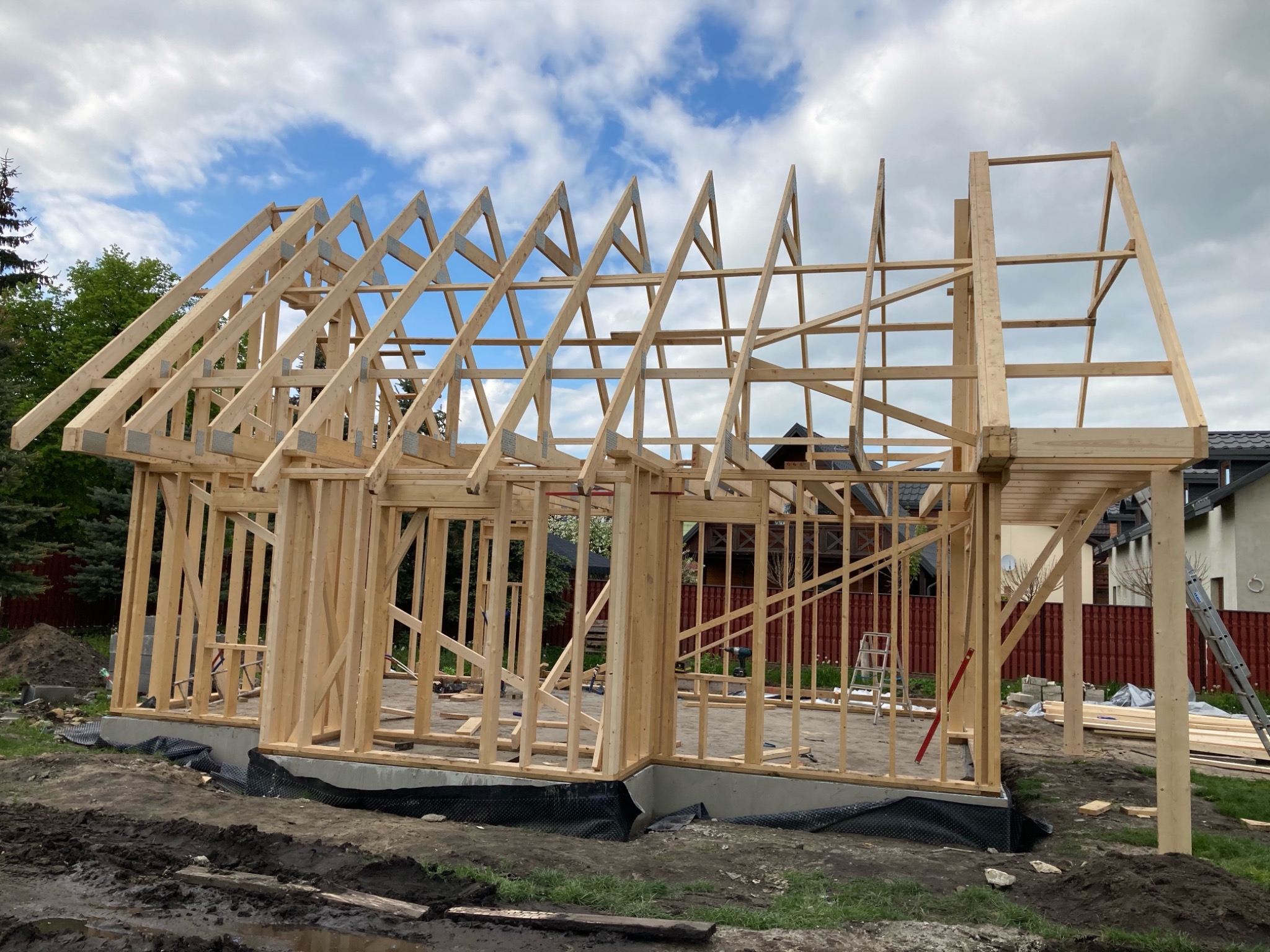 Drewniana konstrukcja szkieletowa domu jednorodzinnego w trakcie budowy, widoczne elementy więźby dachowej na tle błękitnego nieba z chmurami.