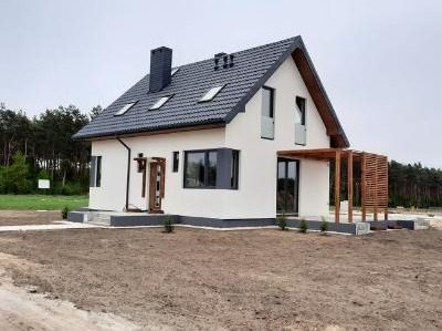 Nowoczesny, dwukondygnacyjny dom z białymi ścianami i ciemnoszarą stolarką okienną oraz dachem z lukarnami, drewnianą pergolą i świeżo zagospodarowanym terenem wokół.