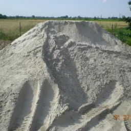 Góra piasku płukanego 0-2mm z wyraźnymi śladami opon pojazdu budowlanego na powierzchni, w tle pole i drzewa.