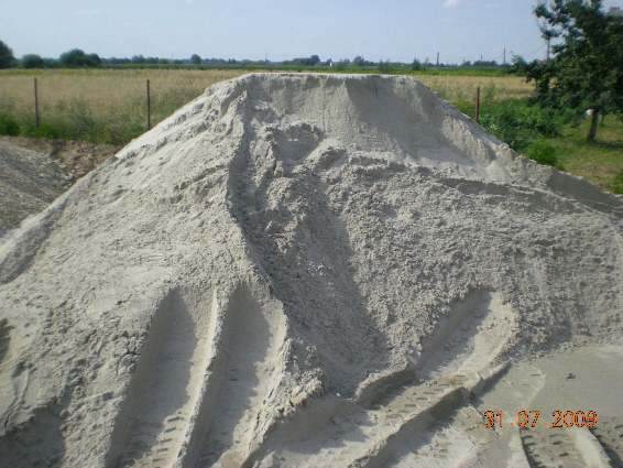 Góra piasku płukanego 0-2mm z wyraźnymi śladami opon pojazdu budowlanego na powierzchni, w tle pole i drzewa.