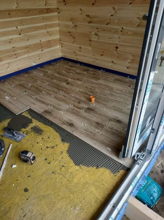 Układanie płytek podłogowych imitujących drewno w pomieszczeniu z boazerią, widoczny klej, poziomica, puszka i krzyżyki dystansowe.