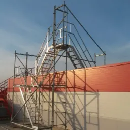 Metalowa konstrukcja rusztowania z drabinkami na tle czerwono-białego budynku w słoneczny dzień.