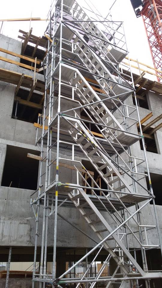 Wysoka konstrukcja rusztowania schodowego, przylegająca do surowej, betonowej ściany budynku w trakcie budowy, częściowo zasłaniająca otwory okienne, z widocznym żurawiem budowlanym w tle.