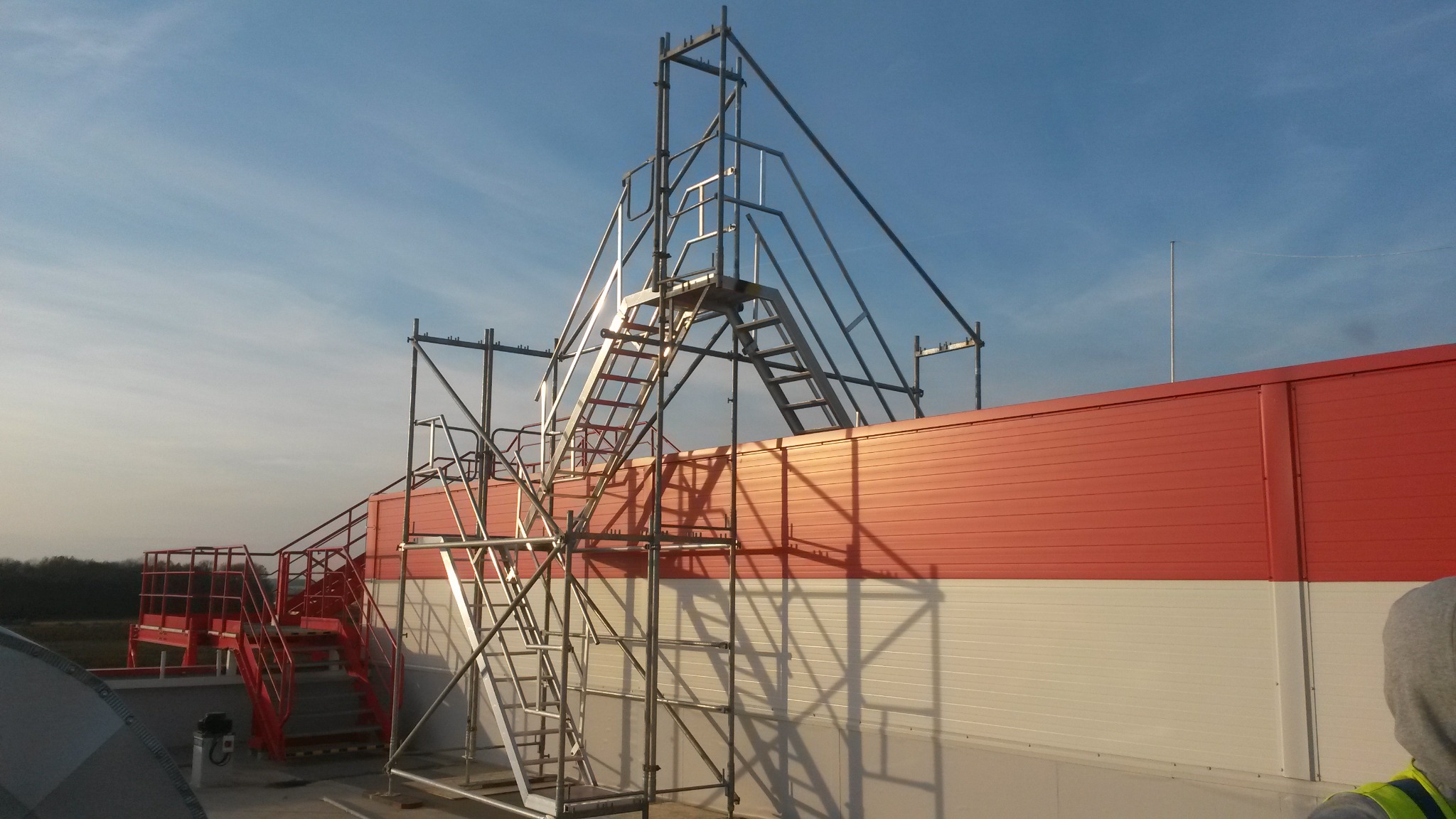 Metalowa konstrukcja rusztowania z drabinkami na tle czerwono-białego budynku w słoneczny dzień.