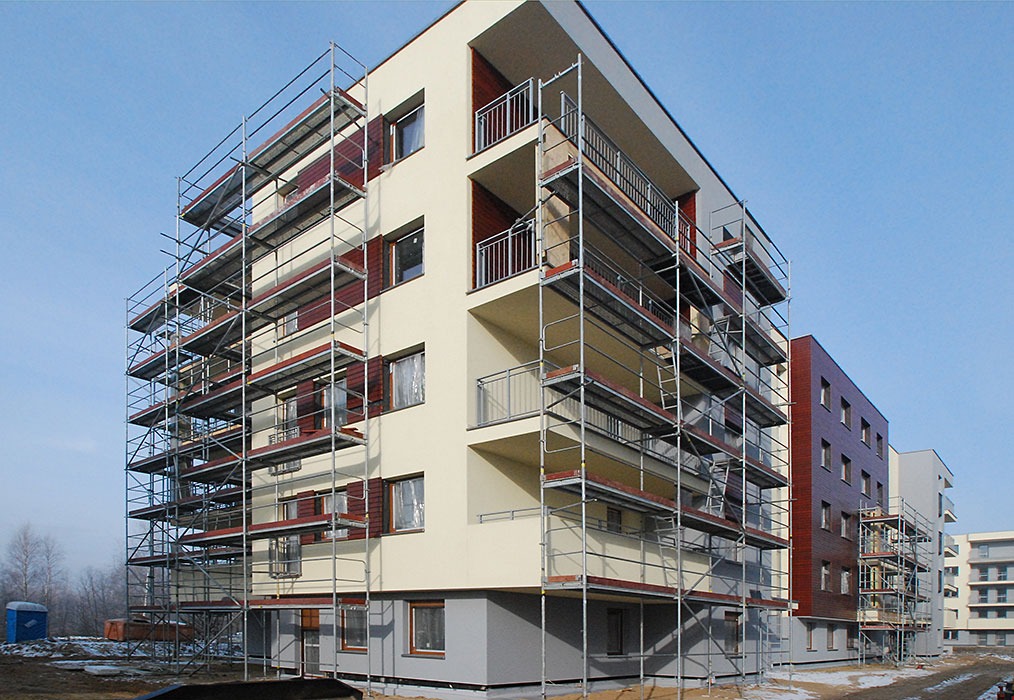 Wielopiętrowy budynek mieszkalny w trakcie prac elewacyjnych, otoczony rusztowaniami, z widocznymi fragmentami elewacji w kolorze beżowym i brązowym.
