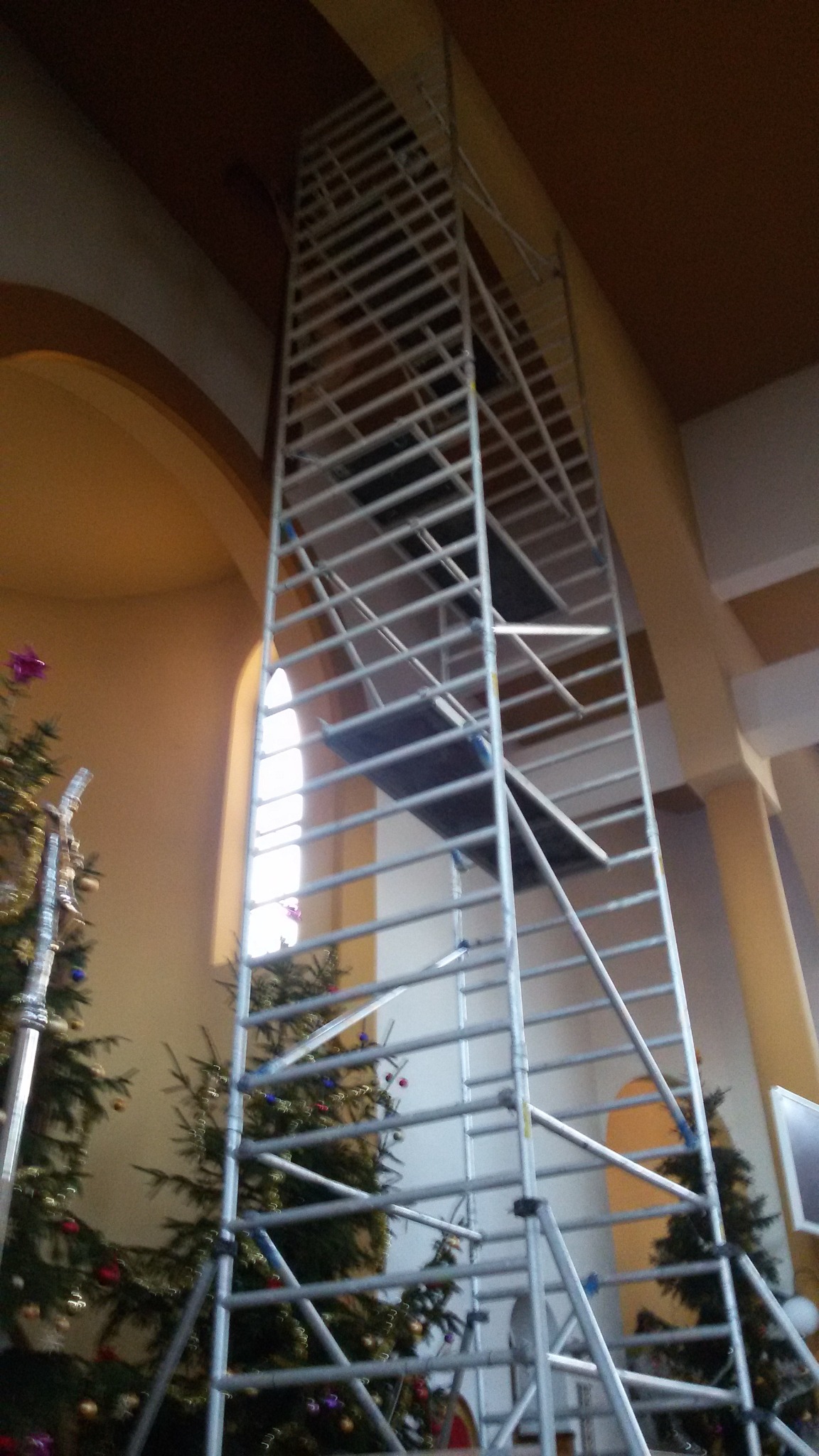 Wysoka aluminiowa wieża rusztowaniowa ustawiona wewnątrz kościoła, widoczne świąteczne drzewka bożonarodzeniowe w tle, oświetlone przez okno. Konstrukcja sięga do sufitu.