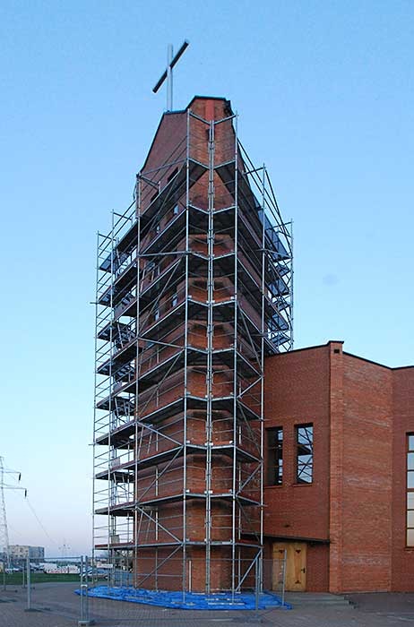 Wieża kościoła z krzyżem na szczycie, otoczona metalowymi rusztowaniami na tle błękitnego nieba, widoczne elementy elewacji z cegły.